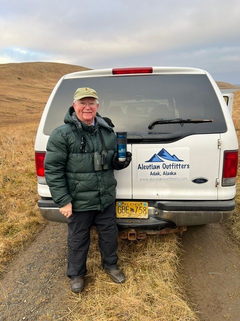 Dr. Gary Rankin stands beside a birding tour vehicle in Alaska wearing binoculars during a wildlife observation trip.