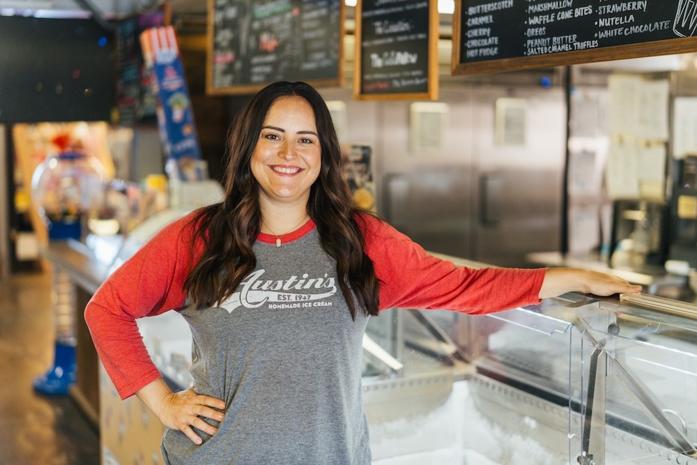 A smiling employee stands behind the counter at Austin’s Ice Cream, with flavor boards and ice cream display cases visible in the background.