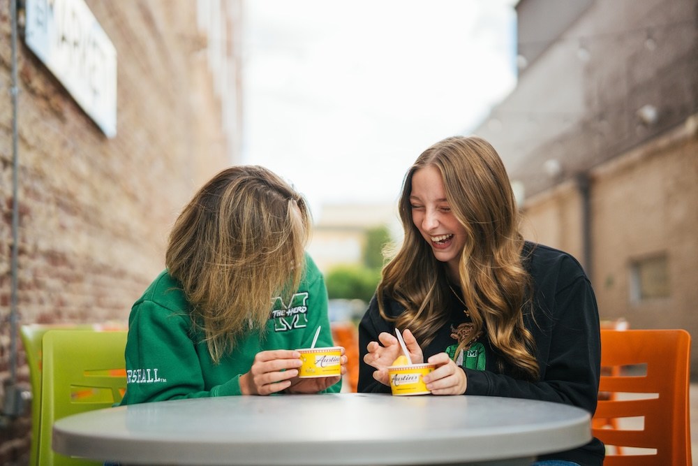 Two students sit at an outdoor table enjoying ice cream from Austin’s Ice Cream, sharing a laugh in a downtown Huntington alleyway seating area.