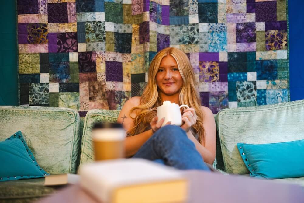 Courtney Burgess relaxes on a couch at Cicada Books, holding a mug in a cozy reading space with a colorful quilt hanging behind her.