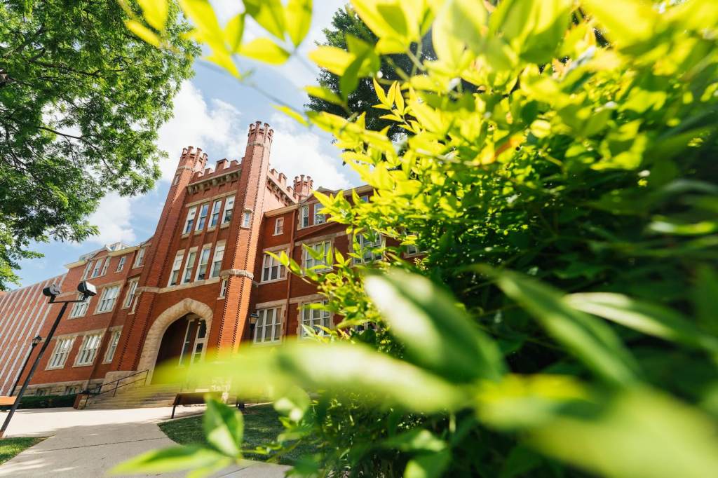 A historic red brick campus building is framed by green leaves on a sunny day.